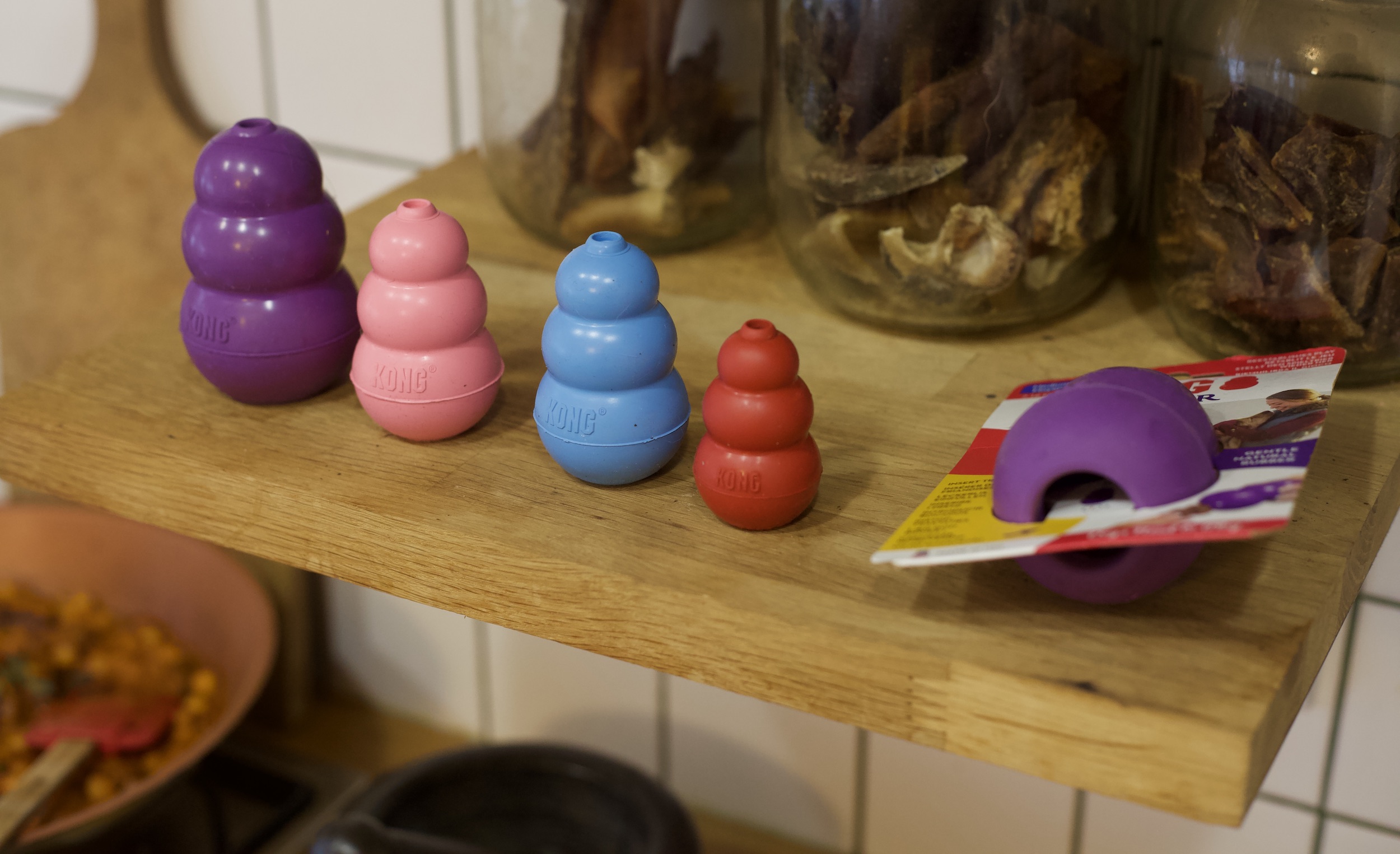 Na kuchennej półce w rzędzie obok siebie stoją cztery zabawki dla psa typu kong. Gumowe gryzaki mają kształt bałwanków, są w różnych rozmiarach i kolorach – fioletowy, różowy, niebieski, czerwony. Ustawione są od największego do najmniejszego.
