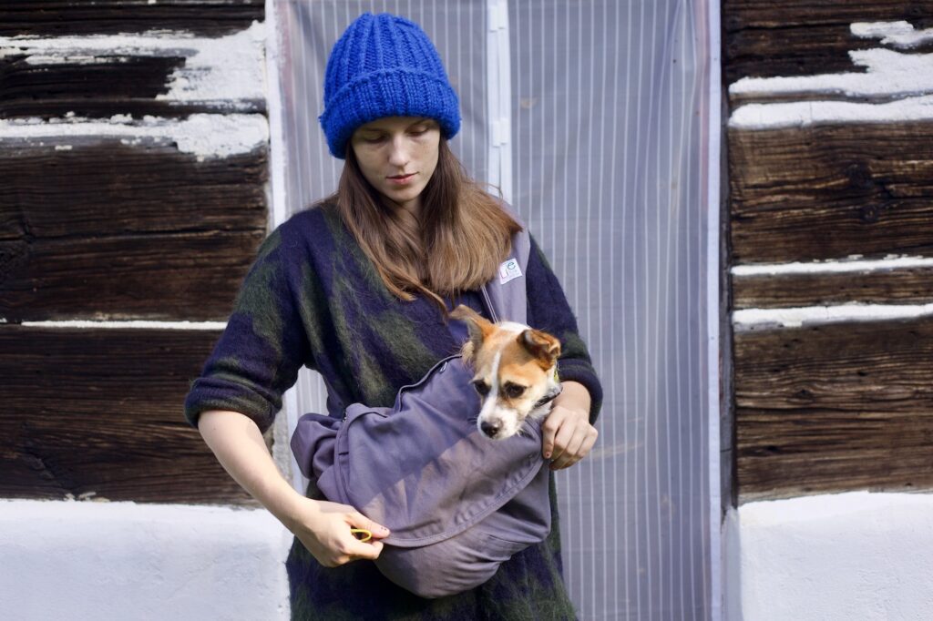 Dziewczyna w swetrze i czapce, z przewieszoną przez ramię szarą torbą, trzyma w środku niej małego pieska. Wystaje mu sama głowa, a ona układa na nim klapę torby.